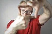 Close-up portrait of a focused individual wearing glasses, framing with hands indoors.
