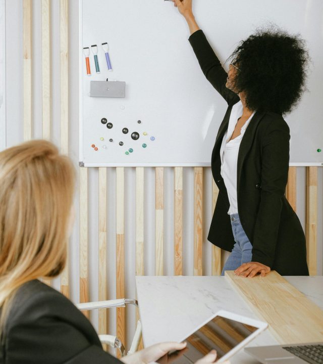 Two women engaged in a business presentation with a whiteboard in a modern office setting.